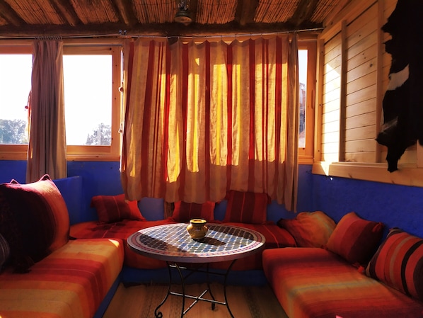 Una habitación acogedora con un sofá azul, una mesa redonda con mantel estampado y una ventana con cortinas a rayas.