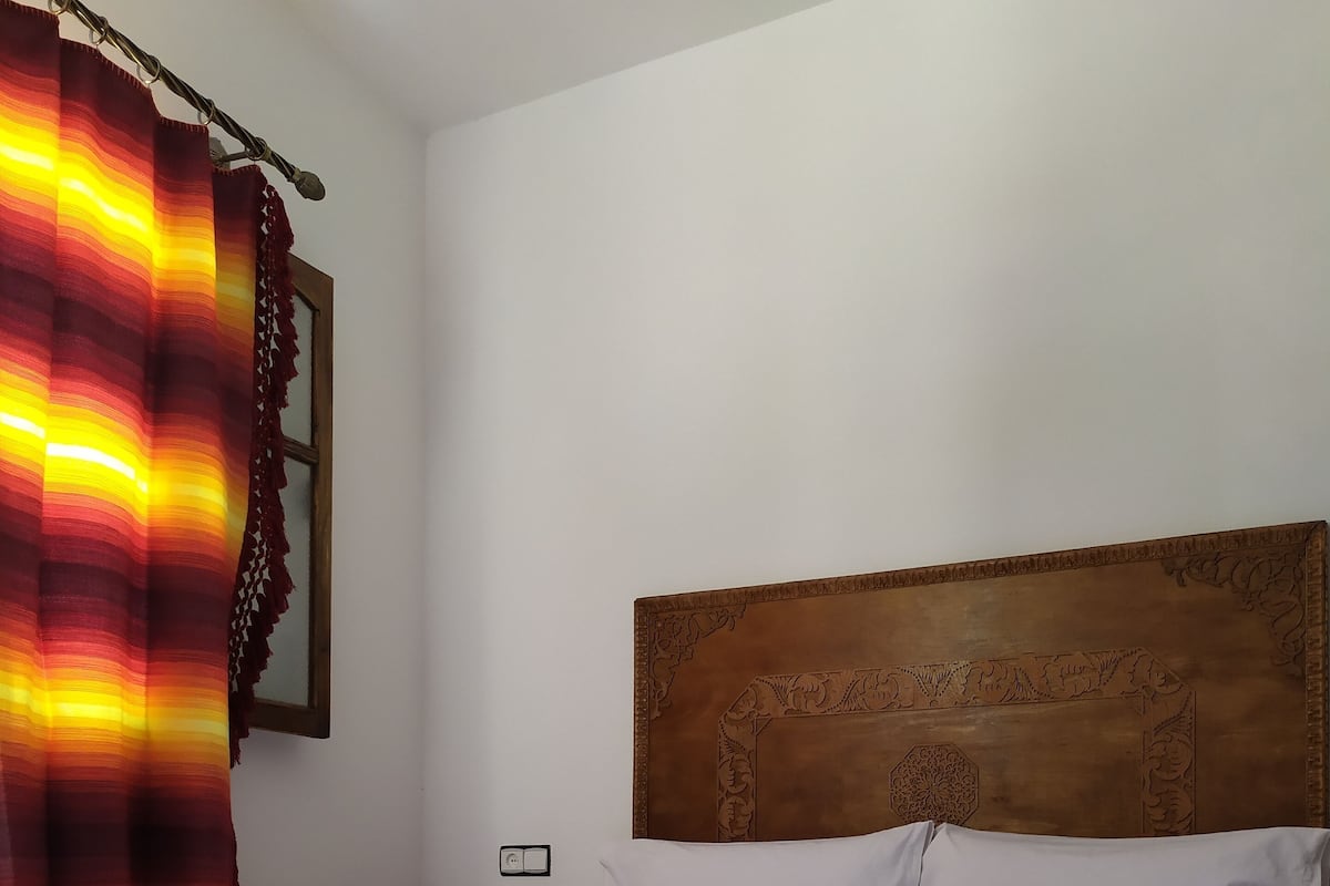 Ein ordentlich bezogenes Bett mit weißen Leinen und gerollten Handtüchern, ein hölzerner Kopfseitenverkleidung mit geschnitzten Details und ein farbenfroher, gestreifter Vorhang.