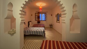 Una habitación con dos camas, piso estampado, una mesita y una ventana con reja decorativa.