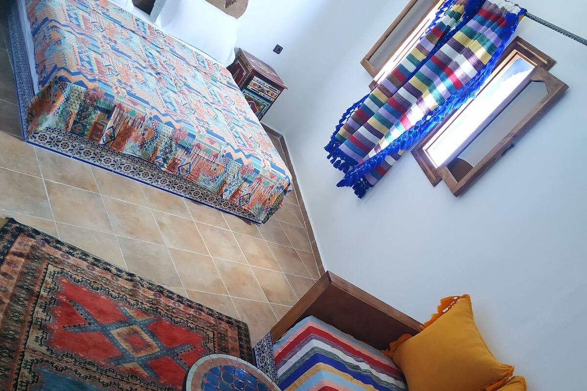 Ein Schlafzimmer mit einem Bett, einem farbenfrohen Teppich, einem gemusterten Teppich, einem kleinen runden Tisch und einem Fenster mit Vorhang.