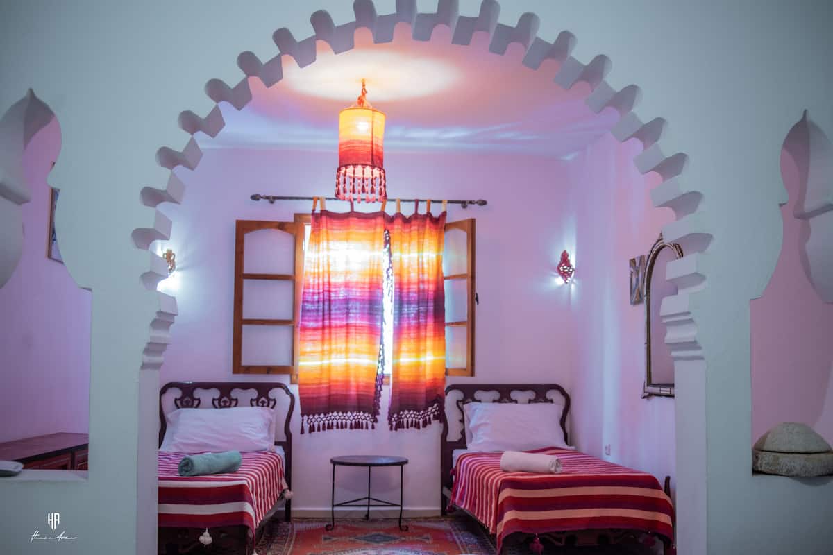 Ein Zimmer mit zwei Betten, einem kleinen Tisch und einem bunten Vorhang.