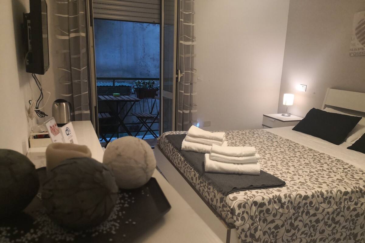 A hotel room with a bed, a desk with a kettle, a small table with chairs, and a view of the outside balcony.
