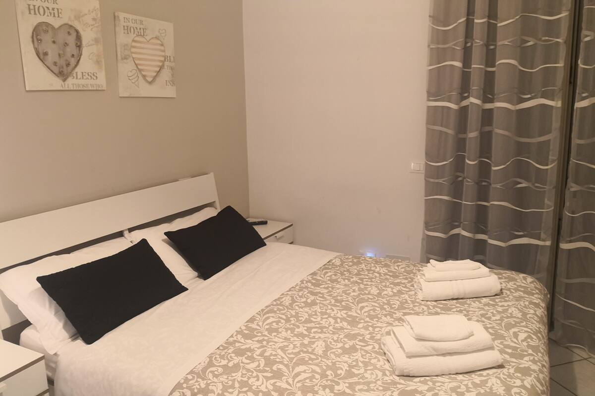 A neatly made bed with a patterned bedspread and two black pillows, a bedside table with a lamp, and two wall art pieces with heart designs.