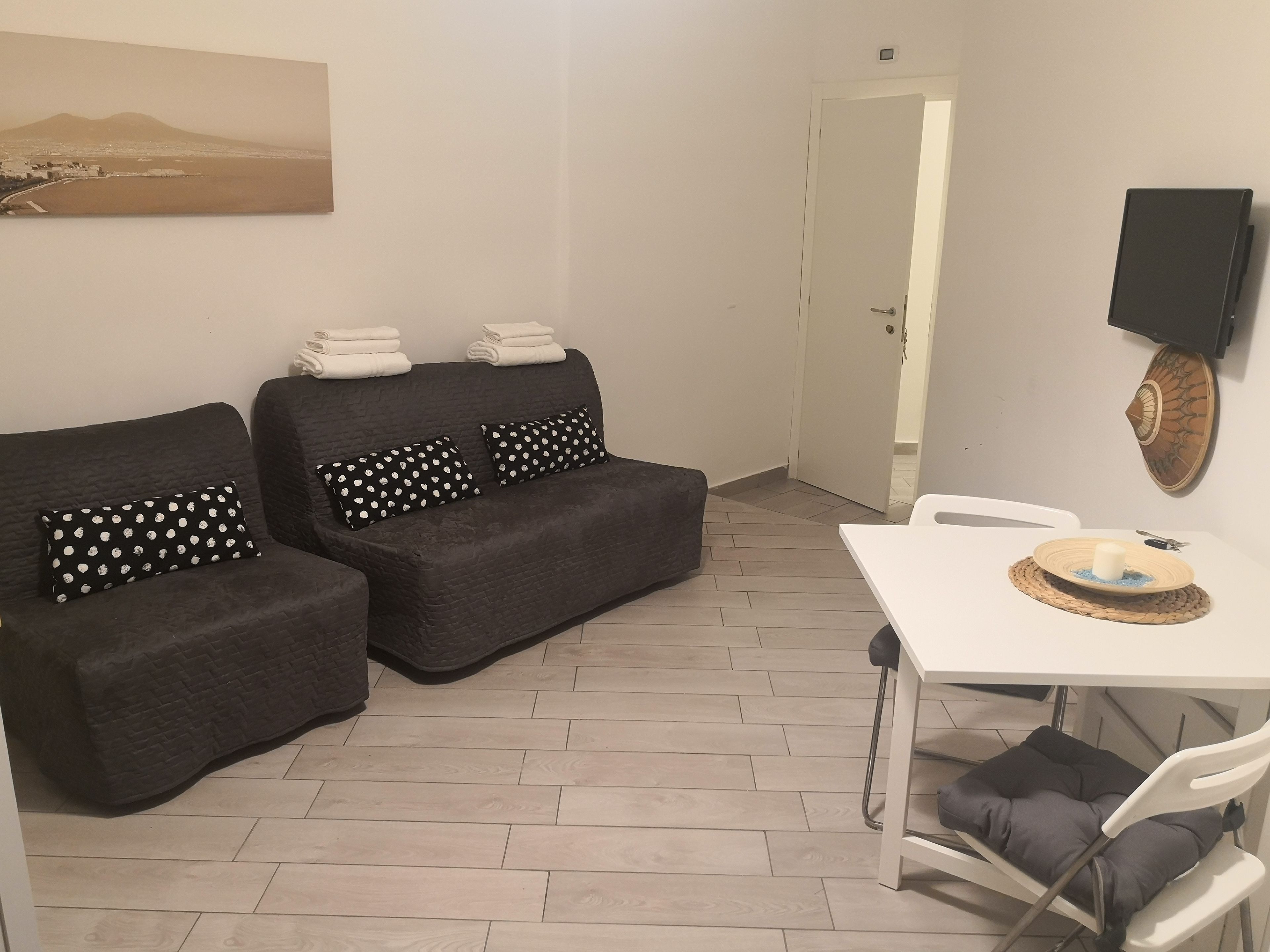 Un salon moderne avec un canapé de couleur gris foncé, deux coussins noirs à pois blancs, une table blanche ornée d’une bougie et d’une assiette décorative, et un téléviseur fixé au mur.