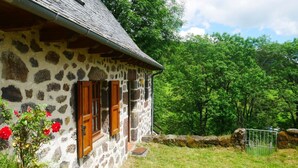 Une maison en pierre avec des volets en bois, un mur en pierre et une cour couverte d’herbe.