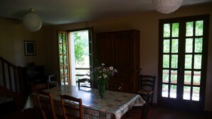 Un coin repas avec une table dressée pour quatre personnes, entourée de chaises en bois. Un buffet en bois se trouve à droite et une bibliothèque à gauche. Un vase rempli de fleurs est posé sur la table.