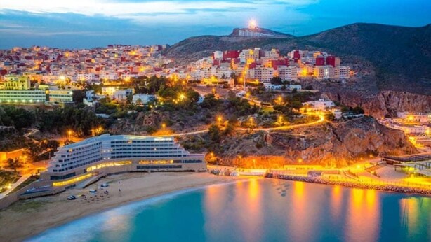 A coastal cityscape at dusk with a prominent hotel featuring a modern design, situated near a beach and a body of water. - pretty apt near beach and cornice (Al Hoceïma)