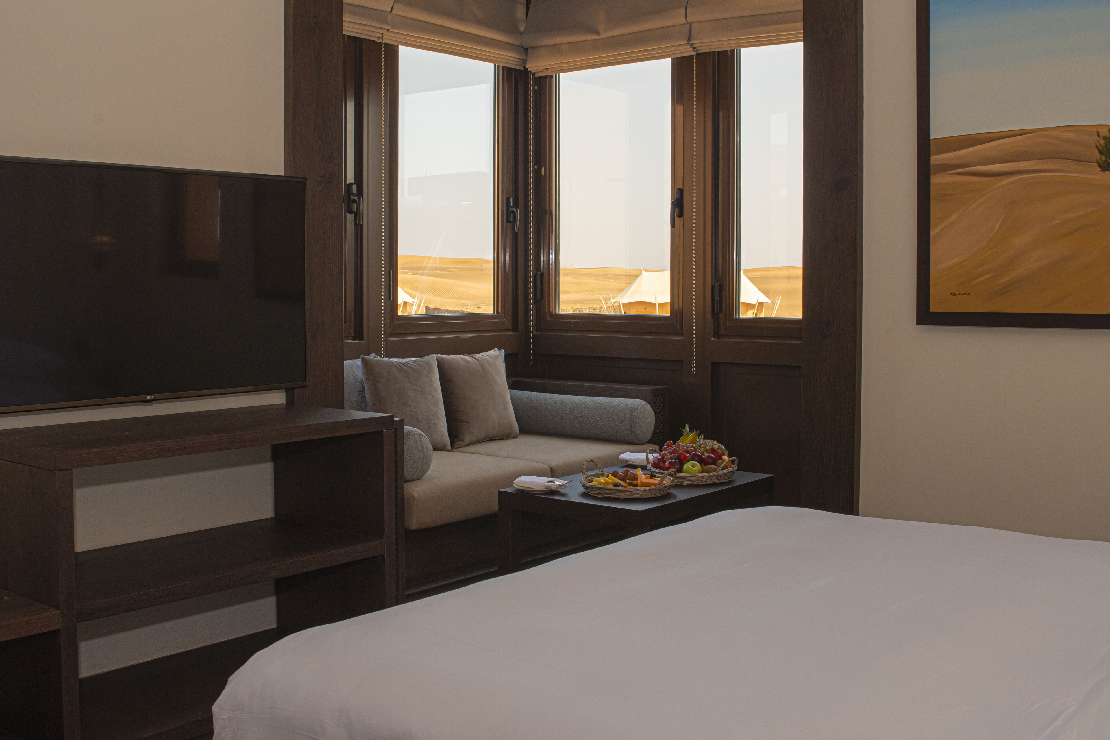 A hotel room with a bed, a sofa, a television, and a dining table with fruit.