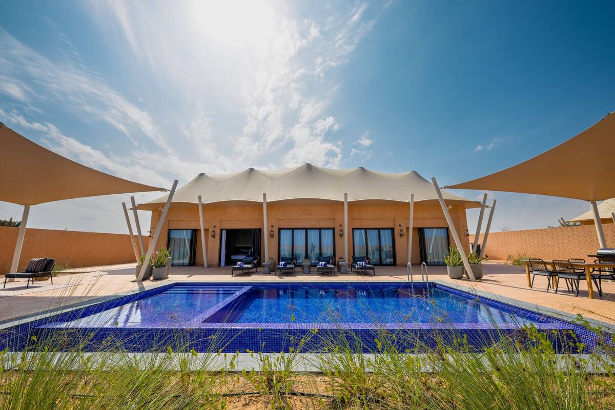 Una piscina con sillones de descanso y sombrillas, un edificio con ventanales grandes, y un cielo despejado.