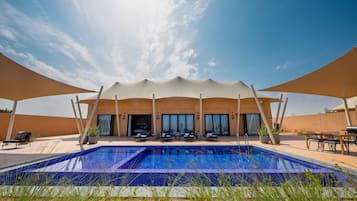 Una piscina con sillones de descanso y sombrillas, un edificio con ventanales grandes, y un cielo despejado.