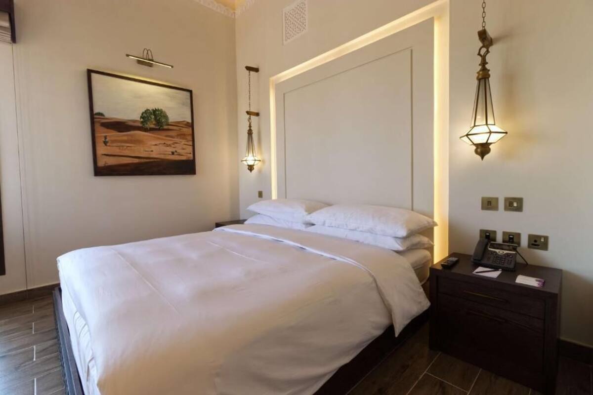 Una cama bien tendida con sábanas blancas en una habitación de hotel, con un cuadro en la pared, una mesita de noche con un teléfono y una lámpara, y un moderno aplique sobre la cama.