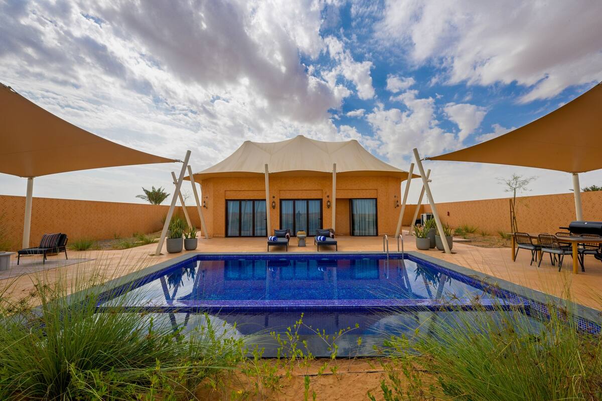 Una piscina con un porche techado, mobiliario exterior y vistas a un edificio con grandes ventanales.