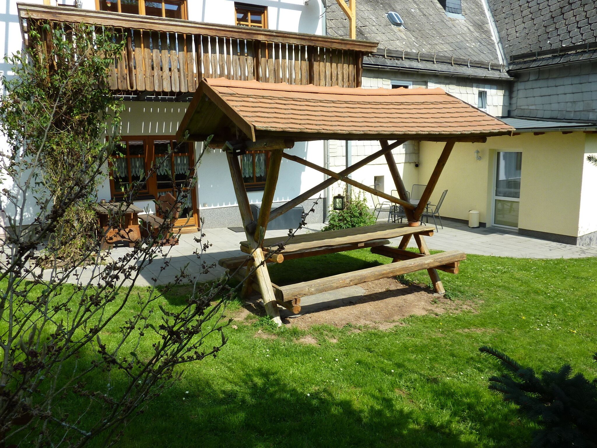 A wooden picnic table with a sloped roof, set on a grassy lawn in front of a building with a white facade and a balcony.