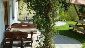 Un espace patio avec une table et des bancs en bois, entouré d’un jardin et d’un bâtiment doté d’un toit en tuiles.
