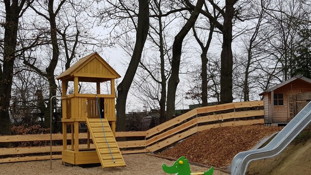 Une aire de jeux avec un toboggan en bois, une structure d’escalade et un petit bâtiment.
