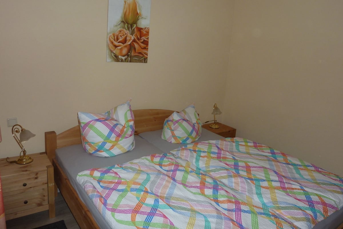 A bedroom with a bed, two bedside tables, a lamp, and a painting of roses on the wall.