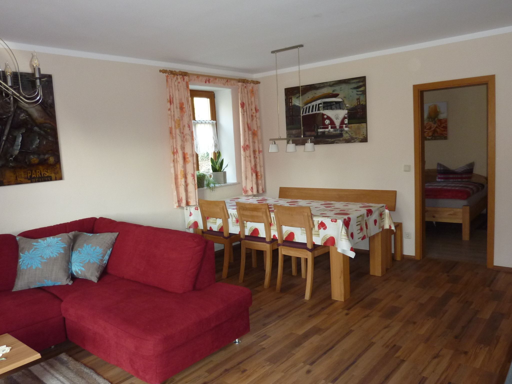 A cozy living room with a red sofa, wooden dining table, and chairs. A painting of a car is on the wall, and there's a window with curtains.