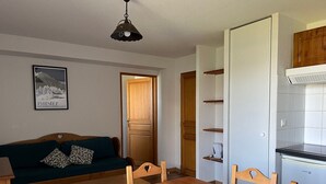 A cozy living room with a wooden sofa, a dining table, and a kitchen area with cabinets and a stove. - Spacious Apt for 6 with Balcony in Peyragudes, 500m from Slopes, Free Spa Access (Peyragudes)