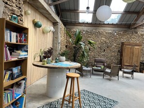 Un coin lecture chaleureux avec une bibliothèque en bois, une table ronde et un tabouret, des chaises en osier et un mur en pierre.
