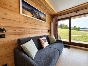 Una sala de estar moderna con pared de madera, un sofá gris con cojines estampados, un cuadro de paisaje enmarco y un amplio ventanal con vistas a un paisaje verde.