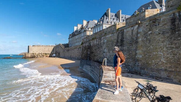 Um passeio costeiro com um muro de pedra, uma praia e um edifício com várias janelas ao fundo.