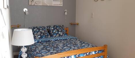 A bedroom with a bed, a bedside table, a lamp, a wall-mounted shelf, and a wall art piece.