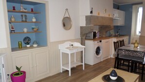 Cozinha compacta com máquina de lavar, área de refeições com mesa e cadeiras, e um pequeno estante com objetos decorativos.