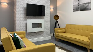 A modern living room with a white brick accent wall, a flat-screen TV, a fireplace, a yellow sofa, and a floor lamp. - GuywoodHouse (Stockport)