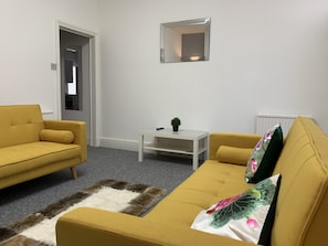 A living room with two yellow sofas, a small white table with a plant, and a mirror on the wall. - GuywoodHouse (Stockport)