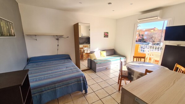 A compact room with a bed, a small table, a chair, a kitchenette, and a window with a view of buildings.