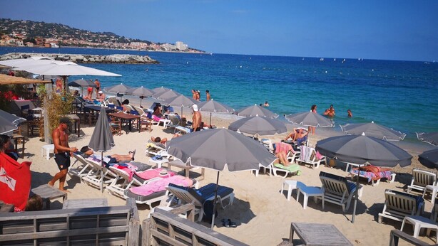 A beach with numerous umbrellas and lounge chairs, a clear view of the sea, and a distant town on the hillside.