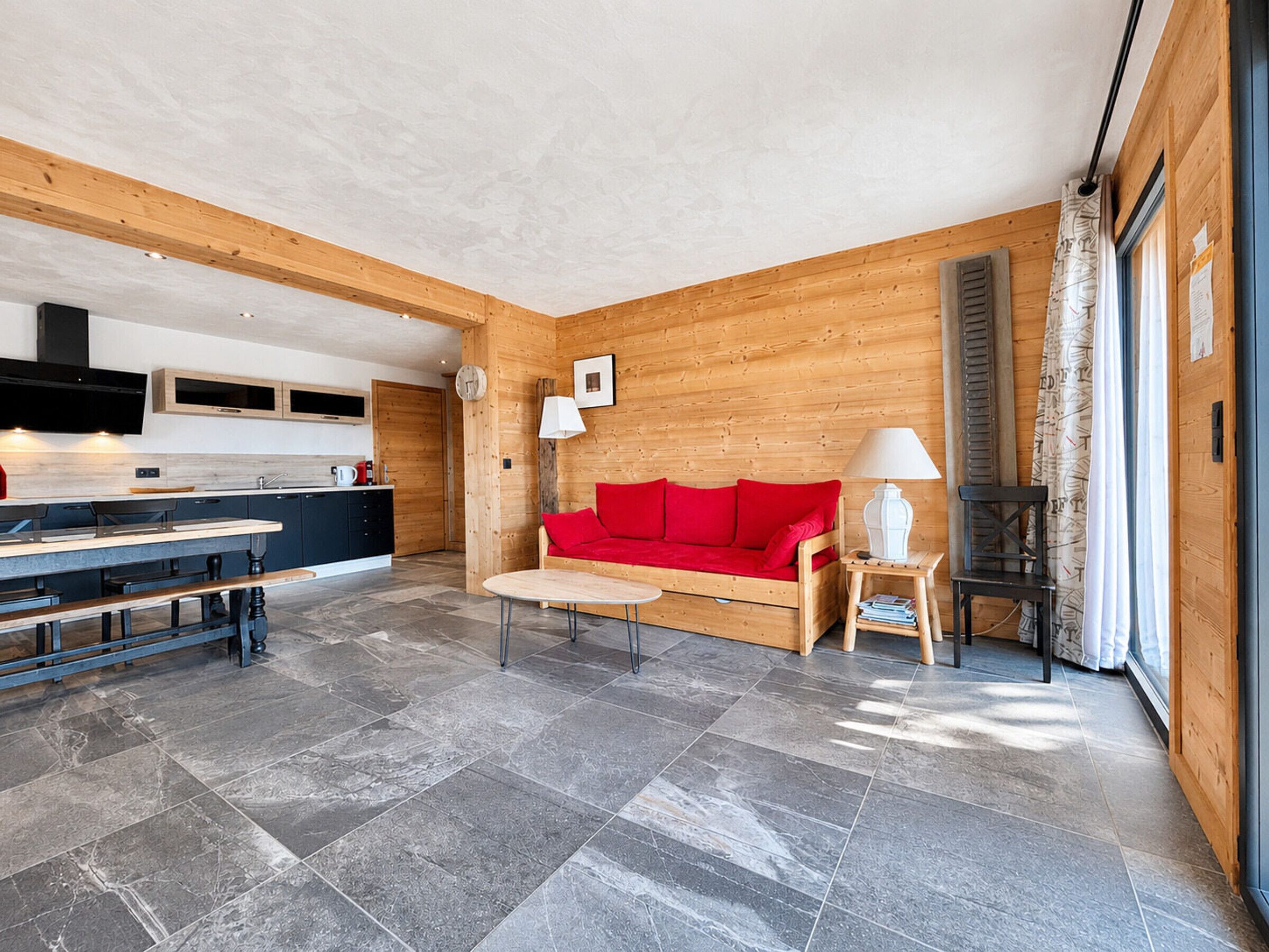Een modern interieur met keuken, eethoek en zithoek, met een rode bank, houten wanden en stenen vloer.