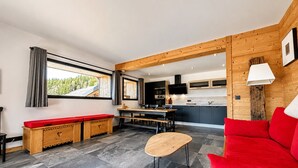 A modern interior with a wooden bench, a red sofa, a small table, and a kitchen area with cabinets and a TV. - Charming apartment with large terrace and parking (Notre-Dame-de-Bellecombe)