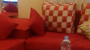 Sala de estar com um sofá vermelho, uma mesa de centro branca com frutas e uma garrafa de água, e uma almofada xadrez.