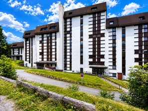 Un immeuble à plusieurs étages, doté d’une façade blanche et de cadres de fenêtres marron foncé, se dressant sur un ciel bleu clair.