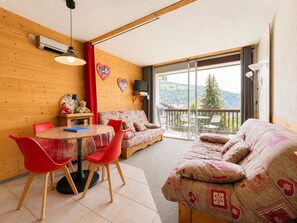 A cozy living room with a wooden floor, a round dining table with red chairs, a sofa with patterned cushions, and a large window offering a view of the outdoors.