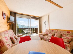 Une pièce avec un plafond en bois, un canapé agrémenté de coussins à motifs, un fauteuil rouge, une table en bois et un téléviseur posé sur un meuble.
