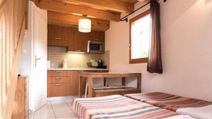 Ein kleines Zimmer mit einer Küchenzeile, zwei Betten mit gestreifter Bettwäsche, einem Holztisch und einem Fenster mit Vorhängen.