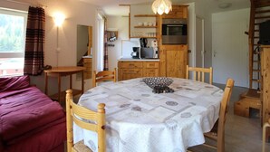 A cozy room with a wooden dining table, chairs, a purple sofa, and a kitchen area with wooden cabinets and appliances.