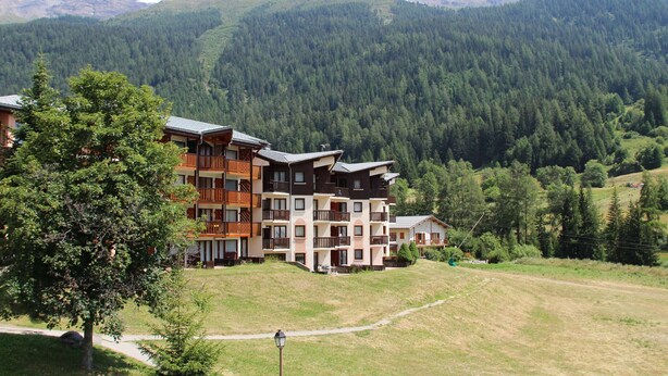 A multi-story hotel with balconies, set against a backdrop of a forested mountain range.
