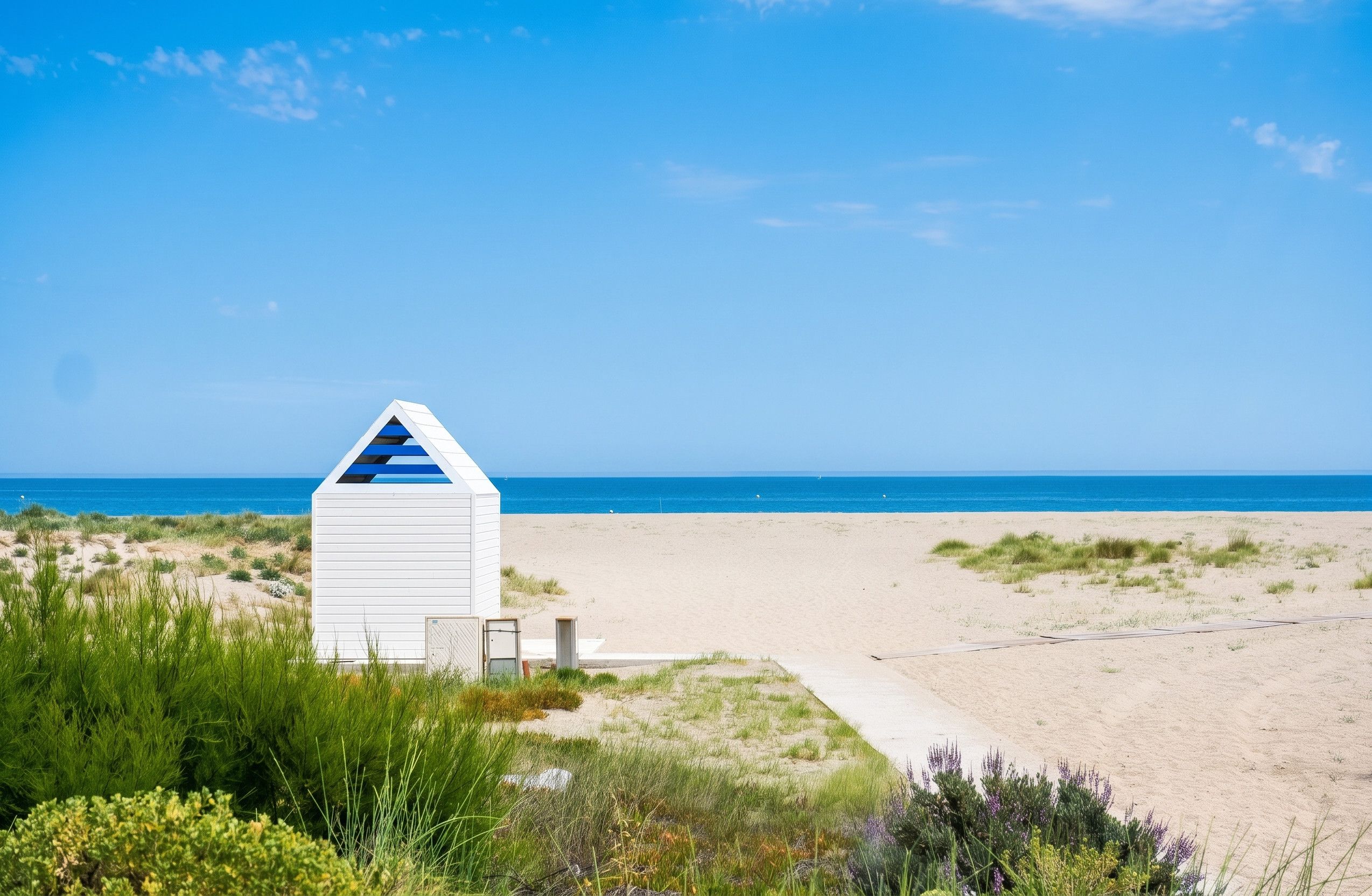 Eine weiße Strandhütte mit geneigtem Dach, gelegen an einem Sandstrand mit Dünen und Vegetation im Vordergrund.