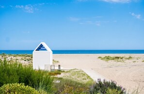 A white beach hut with a sloped roof, situated on a sandy beach with dunes and vegetation in the foreground.
