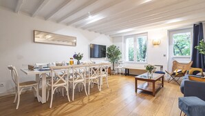 Un vaste espace de vie doté d’un plancher en bois, d’une table à manger avec des chaises, d’une table basse, d’un téléviseur fixé au mur et de grandes fenêtres offrant une vue sur la verdure environnante.