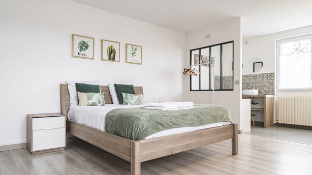Une chambre moderne avec un lit en bois, une literie blanche et un plaid vert. Trois reproductions botaniques encadrées ornent le mur.