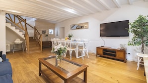 Un vaste salon agrémenté d’un escalier en bois, un coin repas avec des chaises blanches, un téléviseur à écran plat fixé au mur et une grande plante en pot.