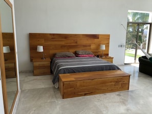 Un dormitorio moderno con cabecera de madera, dos mesitas de noche con lámparas, un banco al pie de la cama y un ventanal con vistas al exterior.