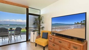 Ein Zimmer mit einem großen Fernseher, der eine Strandansicht zeigt, einem gelben Sessel, einem Korbschrank und einem Essbereich mit Stühlen und Tisch.