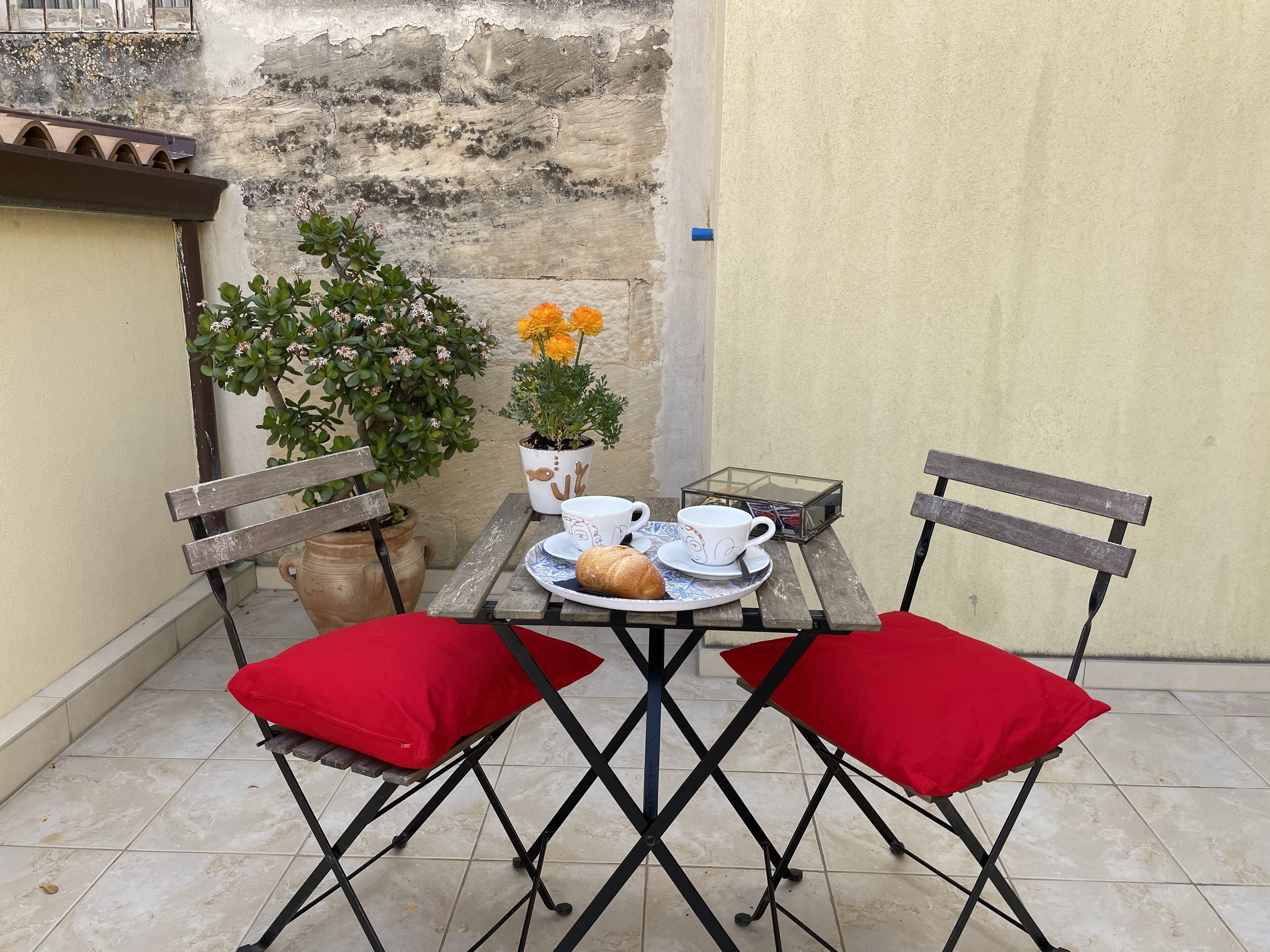 Un pequeño espacio al aire libre con una mesa dispuesta para dos personas, destacando una planta en maceta, una vitrina de vidrio y un croissant.
