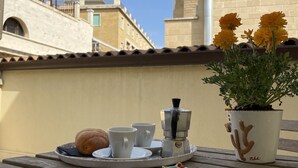 A table with a croissant, two cups, a coffee maker, and a potted plant on a terrace with a tiled floor and a building in the background. - Casa Mamma Elvira (Lecce)