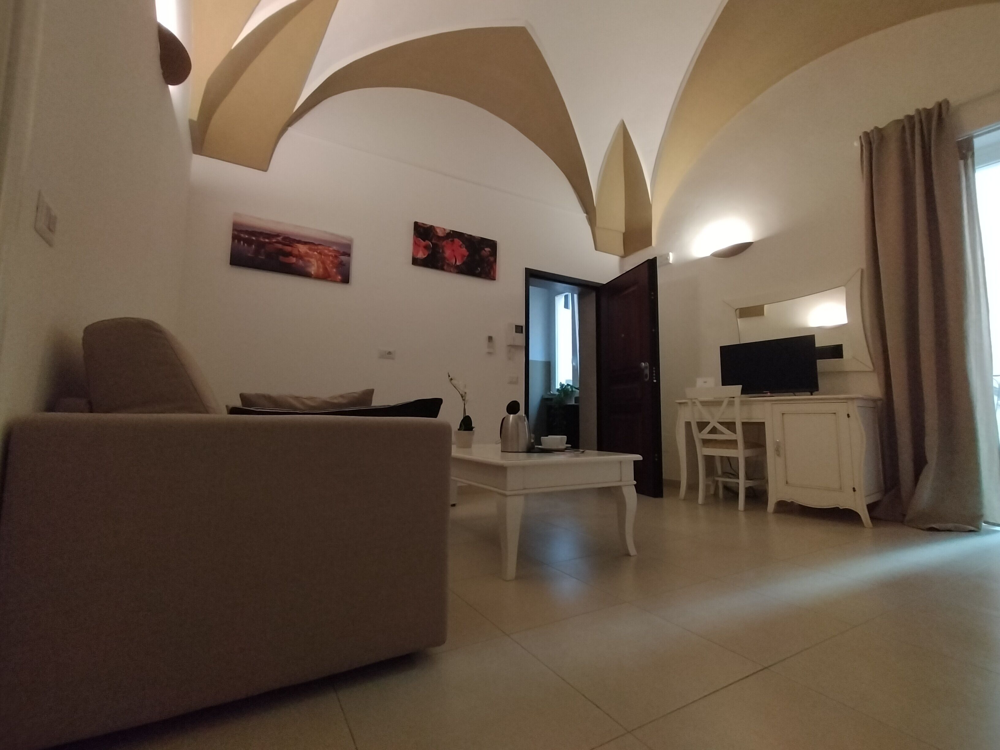 Una sala de estar con un sofá, una mesa de centro, un televisor y un escritorio. Hay dos cuadros en la pared y una puerta que da a otra habitación.